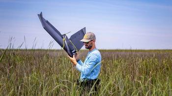 A man carries a drone out of a marsh.